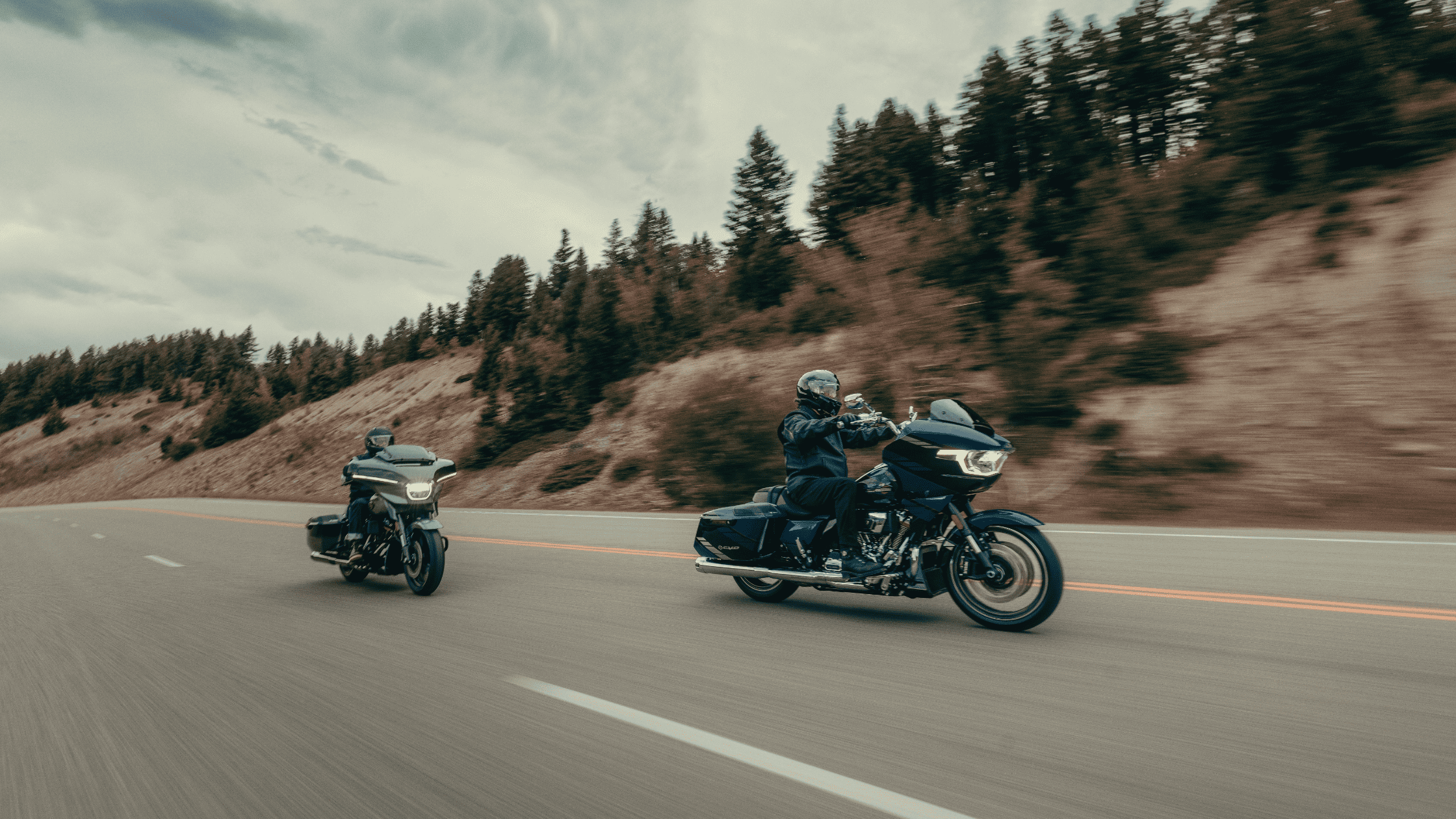 Two riders cruise on Grand American Touring Bikes down a smooth highway surrounded by lush forests.