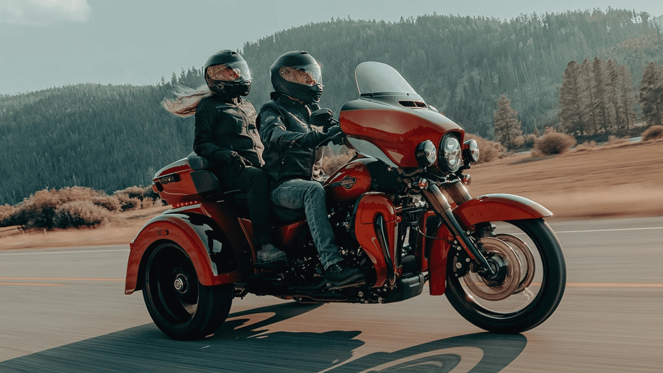 Two riders enjoy a ride on a red Harley-Davidson® Trike cruising down a scenic highway by mountains.