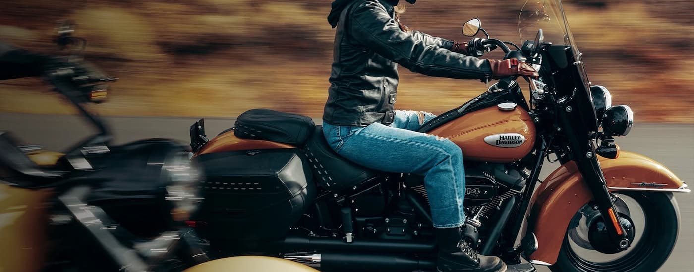 A person is shown riding an orange 2023 Harley-Davidson Heritage Classic from the side on an open …